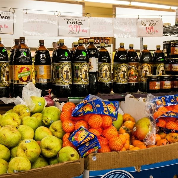 A market stall with bottles of dark beverages, boxes of green pears, and bags of oranges.