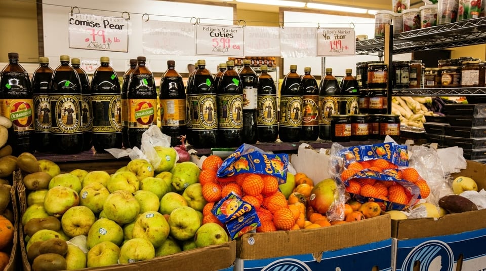 A market stall with bottles of dark beverages, boxes of green pears, and bags of oranges.