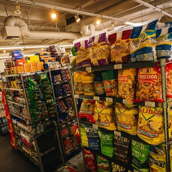 A person pushes a red shopping cart down a grocery store aisle filled with snacks.