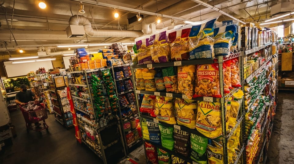 A person pushes a red shopping cart down a grocery store aisle filled with snacks.